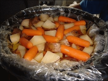 This picture shows bite-sized vegetables on top of beef (either stew meat or pot roast) and onions in a crock pot with a slow cooker liner.