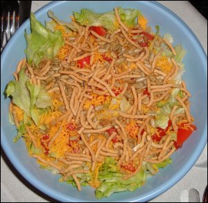 A salad for two with iceberg lettuce, tomatoes, shredded cheddar cheese, chow mein noodles and sunflower kernels.