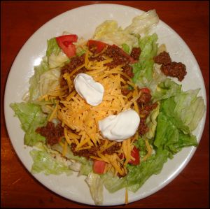 A taco salad with lettuce, tomato, shredded cheddar cheese, sour cream and seasoned taco meat.
