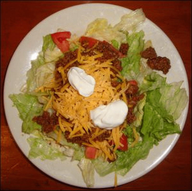 Taco salad, assembled, with corn chips, lettuce, tomato, seasoned taco meat, shredded cheddar cheese and sour cream.