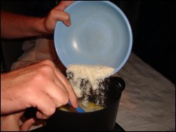 With Vicki holding the hot pot to make sure it does not tip over, Mike pours potato flakes into the water/salt/butter/milk mixture that was prepared according to directions.