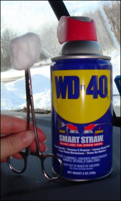 A hemostat holding a cotton ball, next to a can of WD-40.