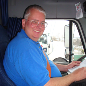 Mike Simons, seated in the driver seat of a semi tractor trailer, is wearing a seat belt.